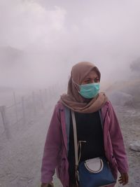 Young woman wearing surgical mask while standing on dirt road in foggy weather