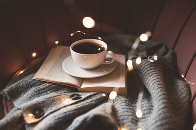 Cup of fresh black tea staying on open paper book with knitted fabric cloth on cozy accent chair 