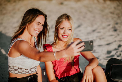 Happy young female friends sitting on sand and taking selfie with mobile phone while spending summer day together on beach