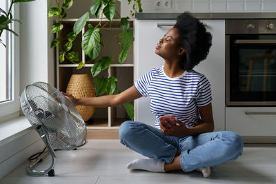 Young african woman sits on floor with phone and catch air currents emanating from electric fan