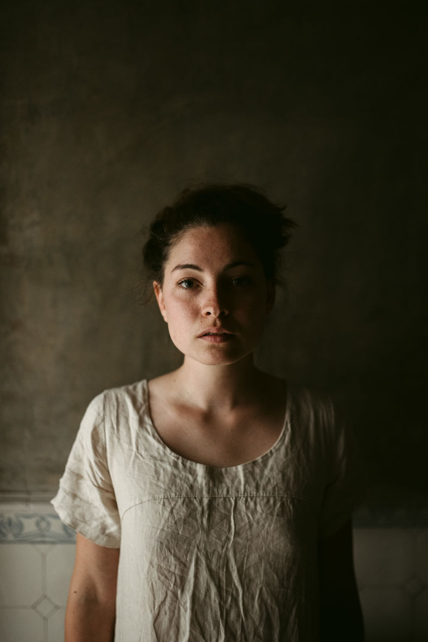 front view, portrait, one person, young adult, looking at camera, indoors, casual clothing, lifestyles, standing, women, real people, young women, waist up, wall - building feature, serious, focus on foreground, adult, hairstyle, contemplation, beautiful woman