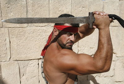 Portrait of shirtless man holding sword against wall