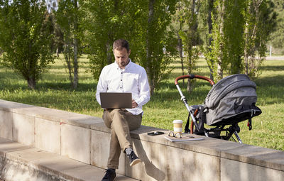 Mid adult businessman working on laptop while sitting by baby stroller in park