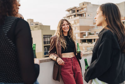 Smiling young woman holding beer bottle and having fun with female friends at rooftop party