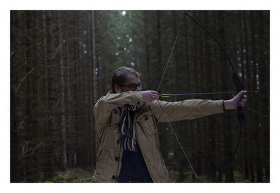 Man holding bow and arrow while standing against trees in forest