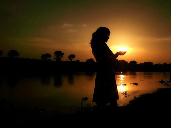 Optical illusion of silhouette woman holding sun by lake against sky during sunset