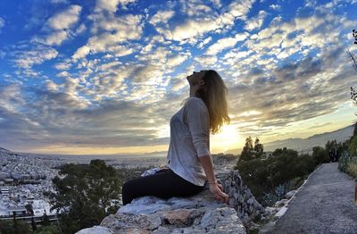 Side view of carefree woman sitting on retaining wall against cloudy sky during sunset