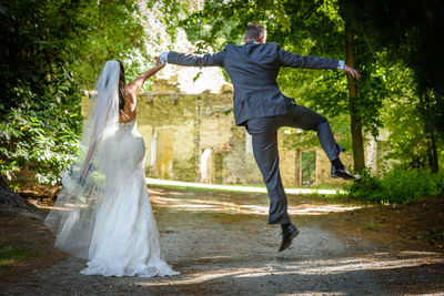 Rear view of groom jumping while holding bride hand on footpath