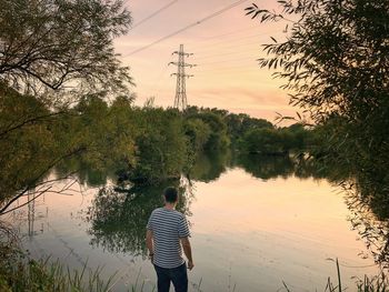 Rear view of man standing by lake against sky during sunset