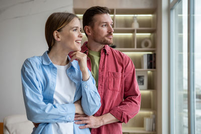 Happy smiling family couple look at window daydreaming waiting guests in housewarming new home.