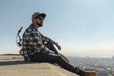 Side view of man wearing sunglasses listening music while sitting on retaining wall against clear blue sky during sunny day