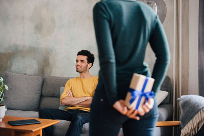 Mid adult man looking at girlfriend hiding gift box while standing in living room