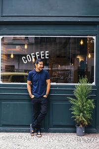 Full length of smiling mature man with hands in pockets standing against coffee shop