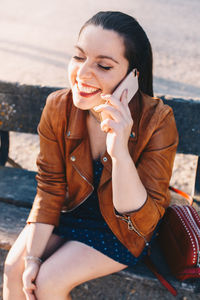 Happy young woman listening over phone while sitting on bench