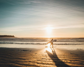 Silhouette man with surfboard walking at beach against sky during sunset