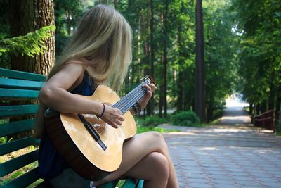 Young woman playing guitar while sitting on bench at park