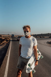 Portrait of man standing on road against clear sky with electric skateboard and mask