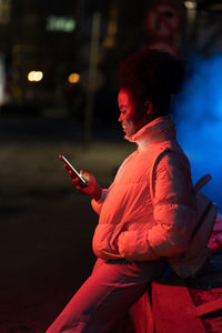 Young african girl chatting online on smartphone while waiting for friend on empty night street