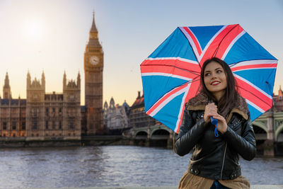 Smiling young woman holding umbrella standing against clock tower in city