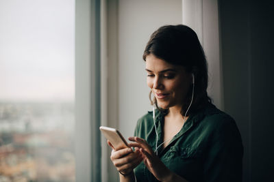 Young woman using phone while standing on window