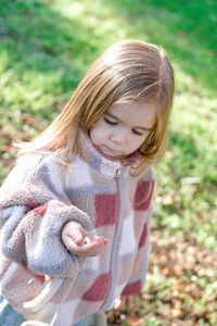 A little girl with long blonde hair in a pink and white checkered jacket kneels in a grassy field. 
