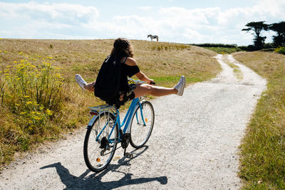 Rear view of carefree woman with legs apart riding bicycle on dirt road during sunny day