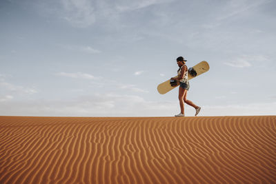 Side view of young female walking on sand and preparing for sandboarding looking away