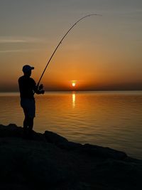 Silhouette man fishing in sea against sky during sunset