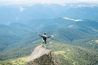 Rear view of man with arms outstretched standing on rock at mountain peak while looking at landscape