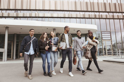 Full length of multi-ethnic university students walking against building in campus