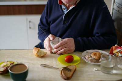 Crop elderly male in casual clothes drinking kefir while sitting at table with bread in light kitchen