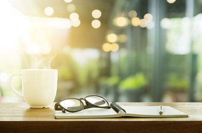 Close-up of coffee cup with diary and eyeglasses on table against window in cafe