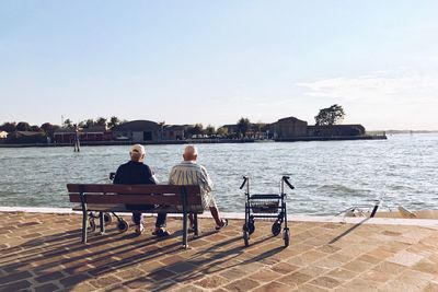 Rear view of friends sitting on bench by lake against sky