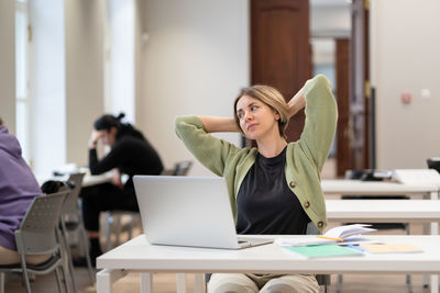 Tired female mature student taking break stretching arms while studying online in university library