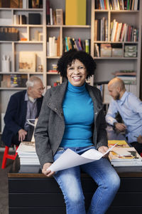 Portrait of happy businesswoman holding blueprint while sitting on desk in office