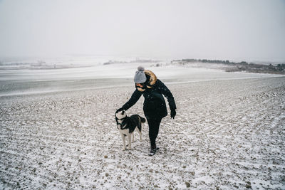 Full body of unrecognizable lady in warm clothes petting obedient husky dog while strolling on snowy field in winter day in countryside under gray cloudless sky