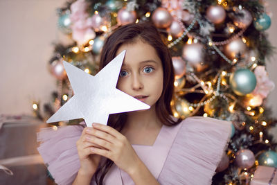 Beautiful baby girl with big eyes in a pink dress is sitting at the christmas tree at home hold star