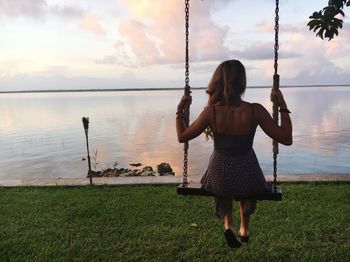 Rear view of woman sitting on swing by sea during sunset