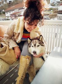 Smiling woman with husky sitting by fence against houses