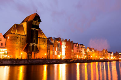 Illuminated buildings by river against sky in old town at dusk