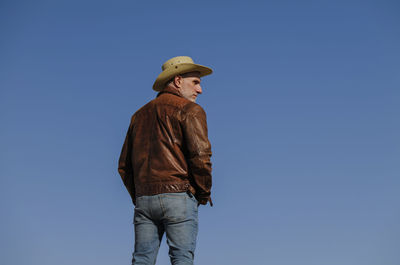 Rear view of adult man with brown leather jacket and cowboy hat against blue clear sky