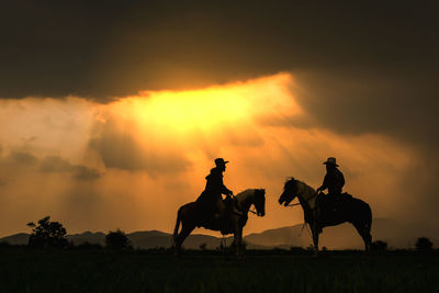 Silhouette cowboys riding horses against cloudy sky during sunset