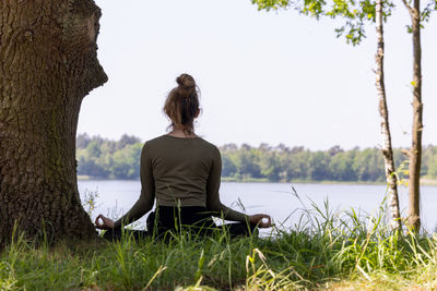 Young woman practicing yoga in nature. sitting on green grass next to a tree and a forest lake. 