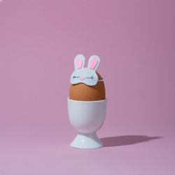 Brown egg in bunny sleep mask in white egg cup on pink background. easter egg concept.