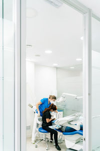 Concentrated female stomatologist in mask sitting near dental chair while treating client with help of nurse in light cabinet