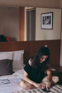 Smiling young woman using mobile phone while sitting on bed at home