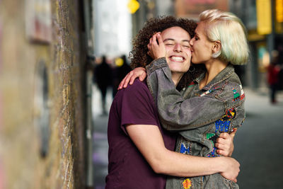 Smiling young woman kissing friend on footpath in city