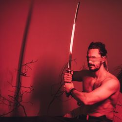 Shirtless man with blindfolds holding sword in illuminated room