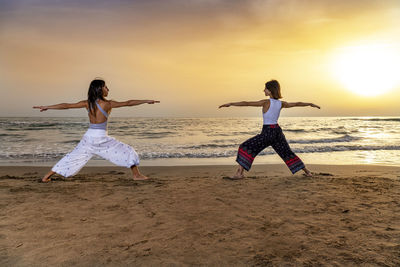 Two young women doing the warrior yoga pose on the beach at sunset. sense of energy and stability.