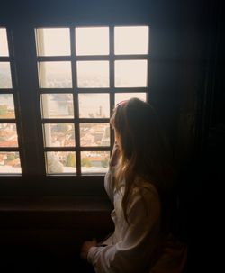 Side view of young woman looking through window at home